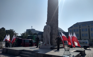 Na zdjęciu widzimy dwóch żołnierzy  i pomnik żołnierza polskiego, w tle budynki, zaparkowane auta i flagę biało-czerwoną